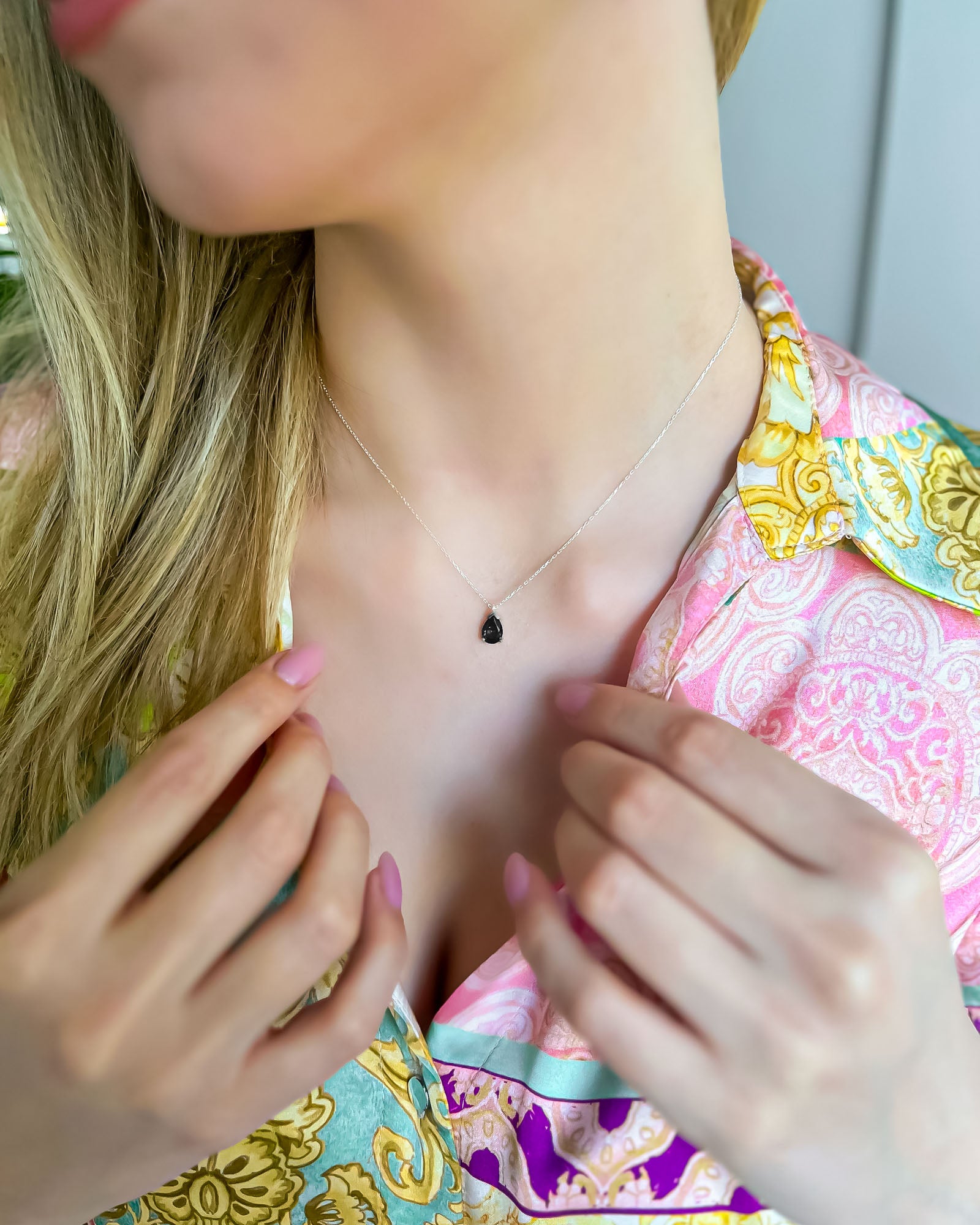 Person wearing a delicate necklace with a small black pendant, set against a colorful patterned fabric background.