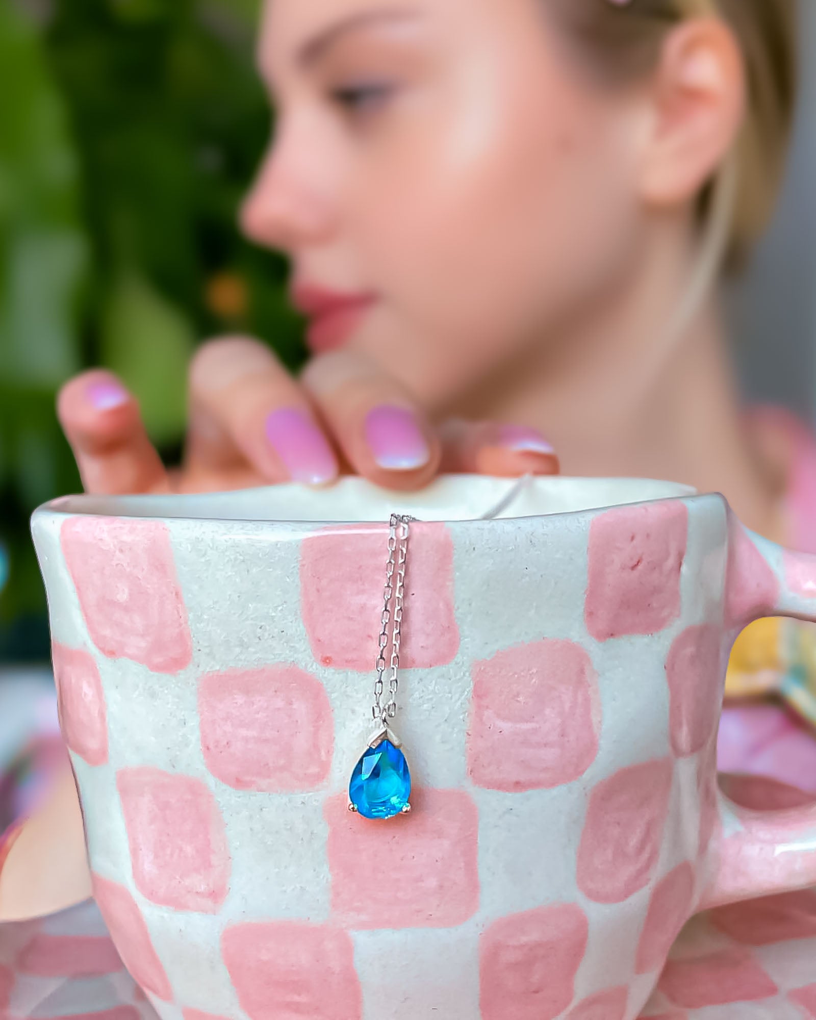 Necklace with a blue teardrop pendant held over a pink checkered mug.