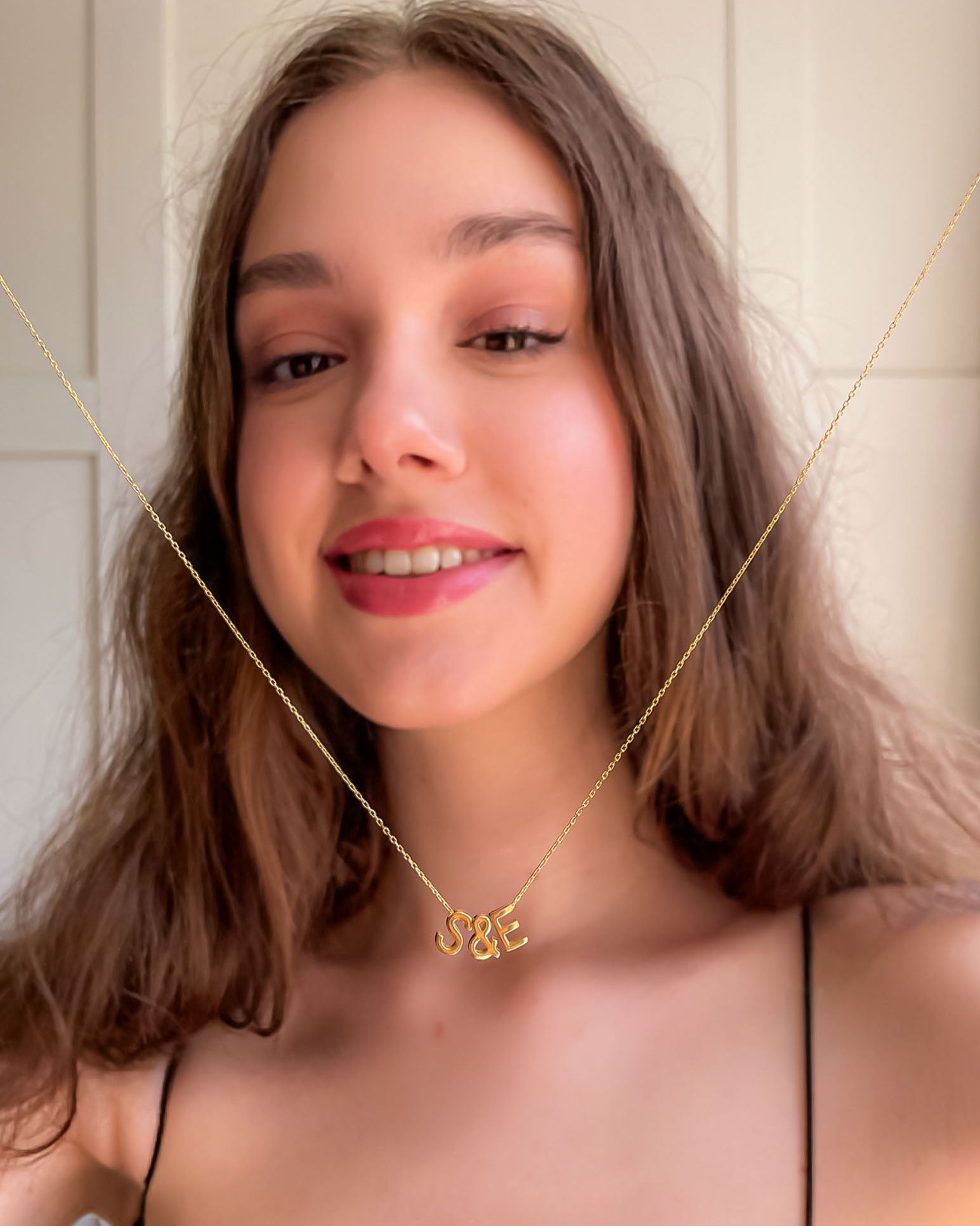 Woman wearing a gold necklace with a 'S&K' pendant against a neutral background